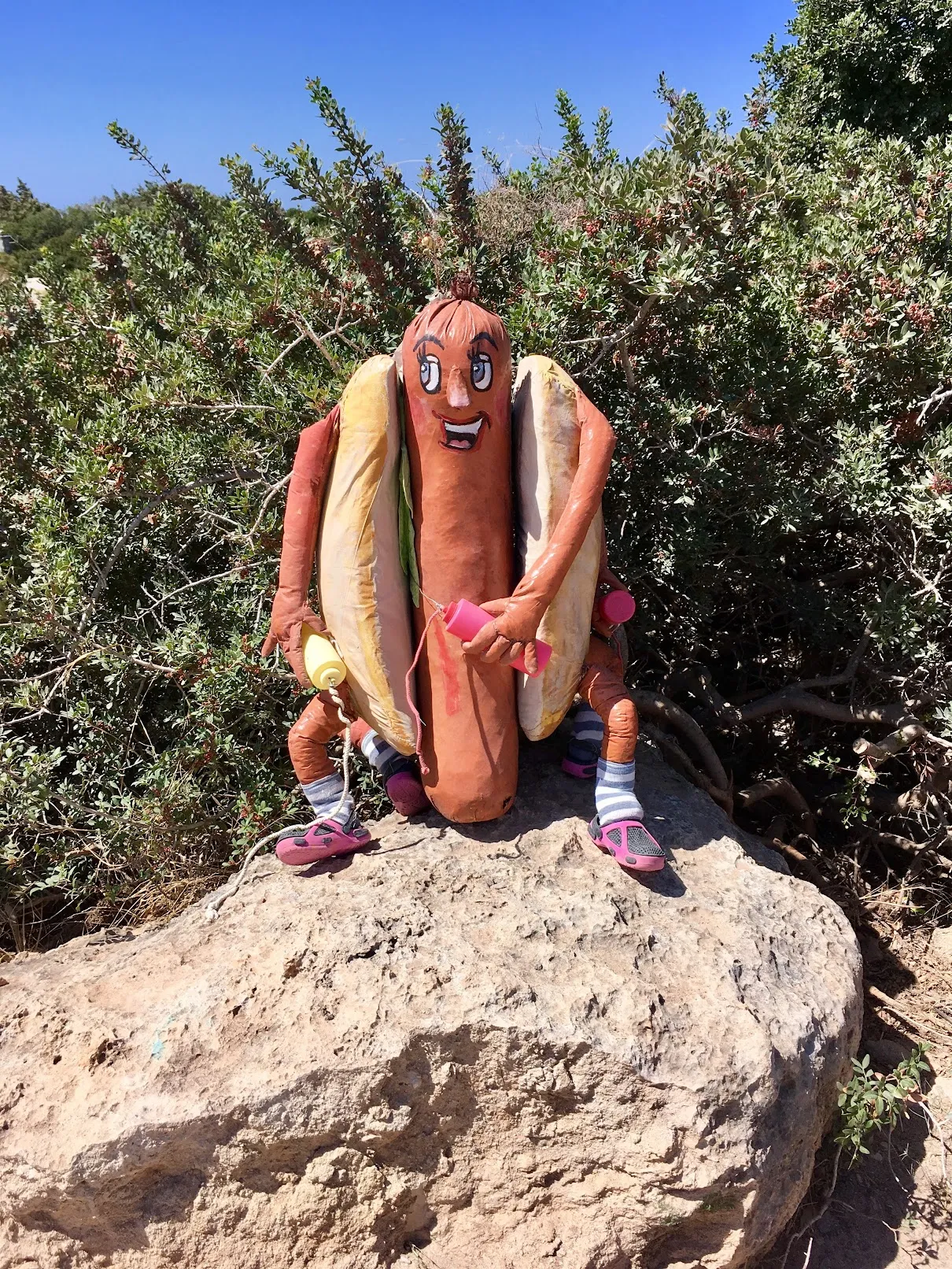 Statue of a smiling hot dog character with arms, legs, and a face, sitting on a rock.