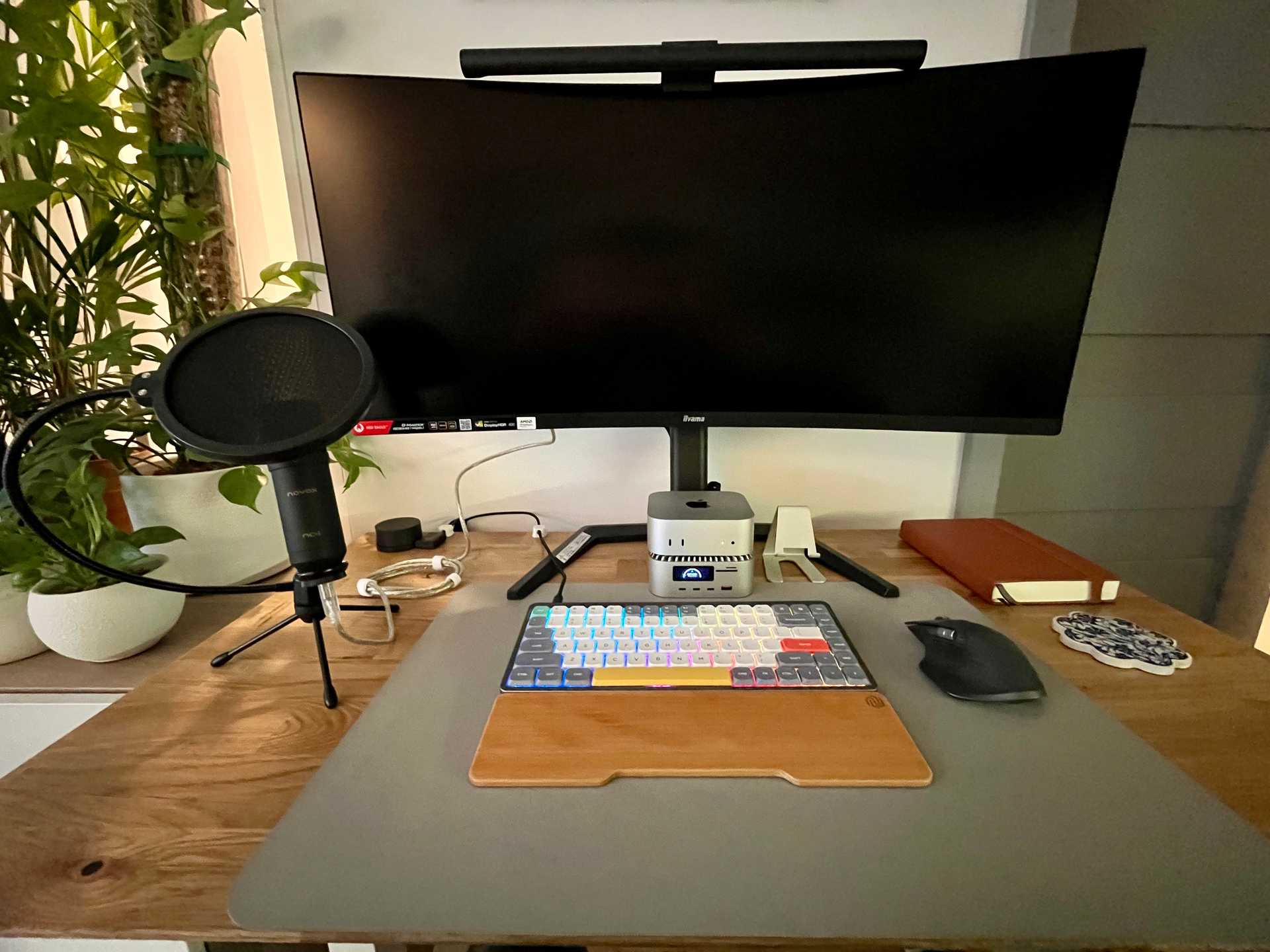 Home office workspace featuring a Computer: Mac mini 2024 (Apple M4, 16 GB RAM, 256 GB SSD) mounted in a RayCue ALL-in-One Charger Dock for Mac mini M4, connected to an iiyama G-Master GCB3481WQSU-B1 34″ UWQHD 180 Hz Monitor with a Xiaomi Mi Computer Monitor Light Bar. On the desk are a NuPhy Air75 Keyboard with Gateron G Pro Red switches, a Logitech MX Master 3S Mouse (Graphite), and a Novox NC-1 USB microphone on a stand. The setup includes a UGREEN DP 1.4 KVM Switch, UGREEN USB-C to DisplayPort 1.4 cables, Thunderbolt 5 USB-C cables, and is placed on an Oak Rustic Desktop mounted on a Silver Monkey EDF-180 desk frame. Part of the workspace ecosystem includes a Synology DiskStation DS423+ NAS with WD Red Plus and WD Red SN700 storage, and a Brother HL-L2352DW printer.